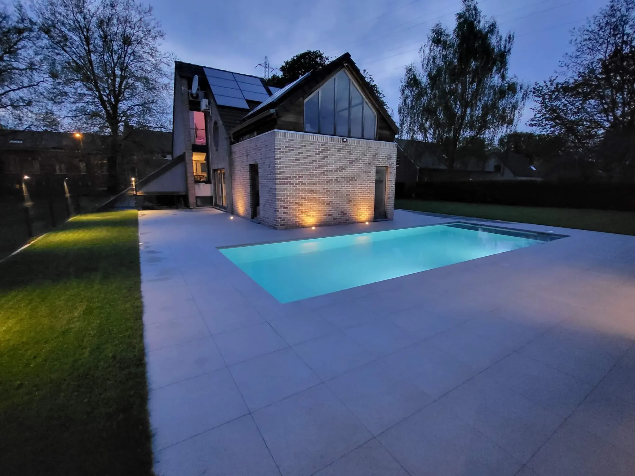 Piscine contemporaine éclairée au crépuscule devant une maison en briques