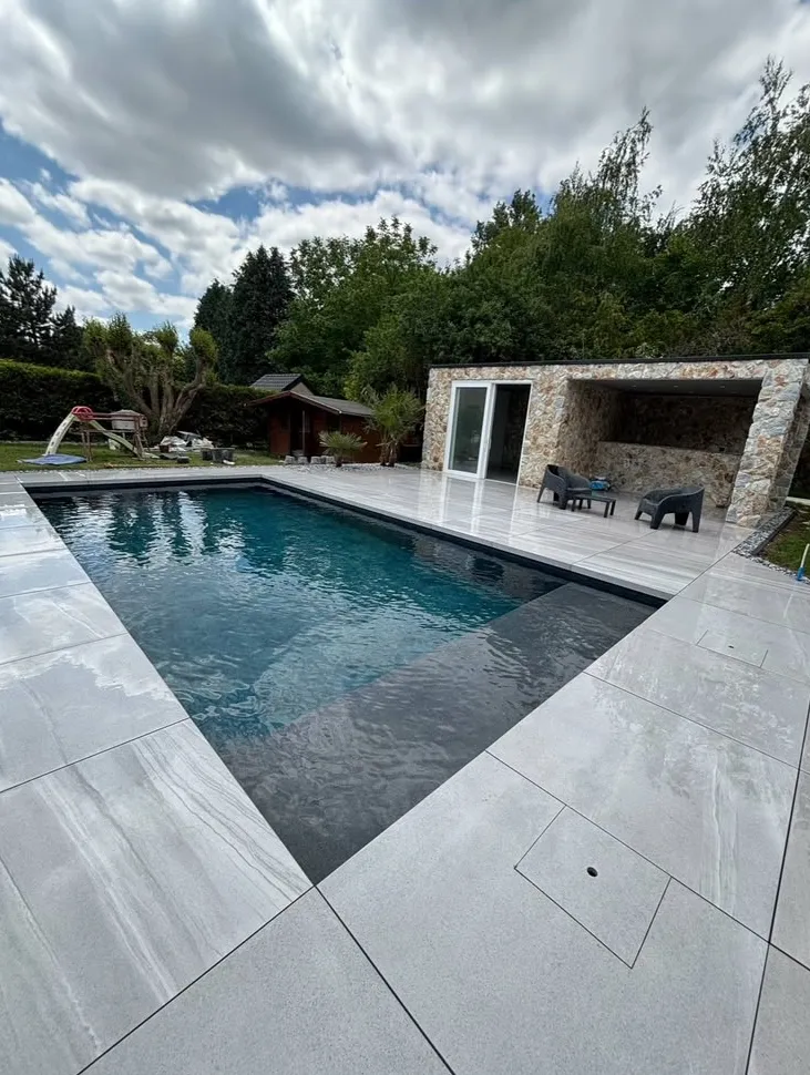 Piscine sombre entourée d’une terrasse claire devant un pool house en pierre