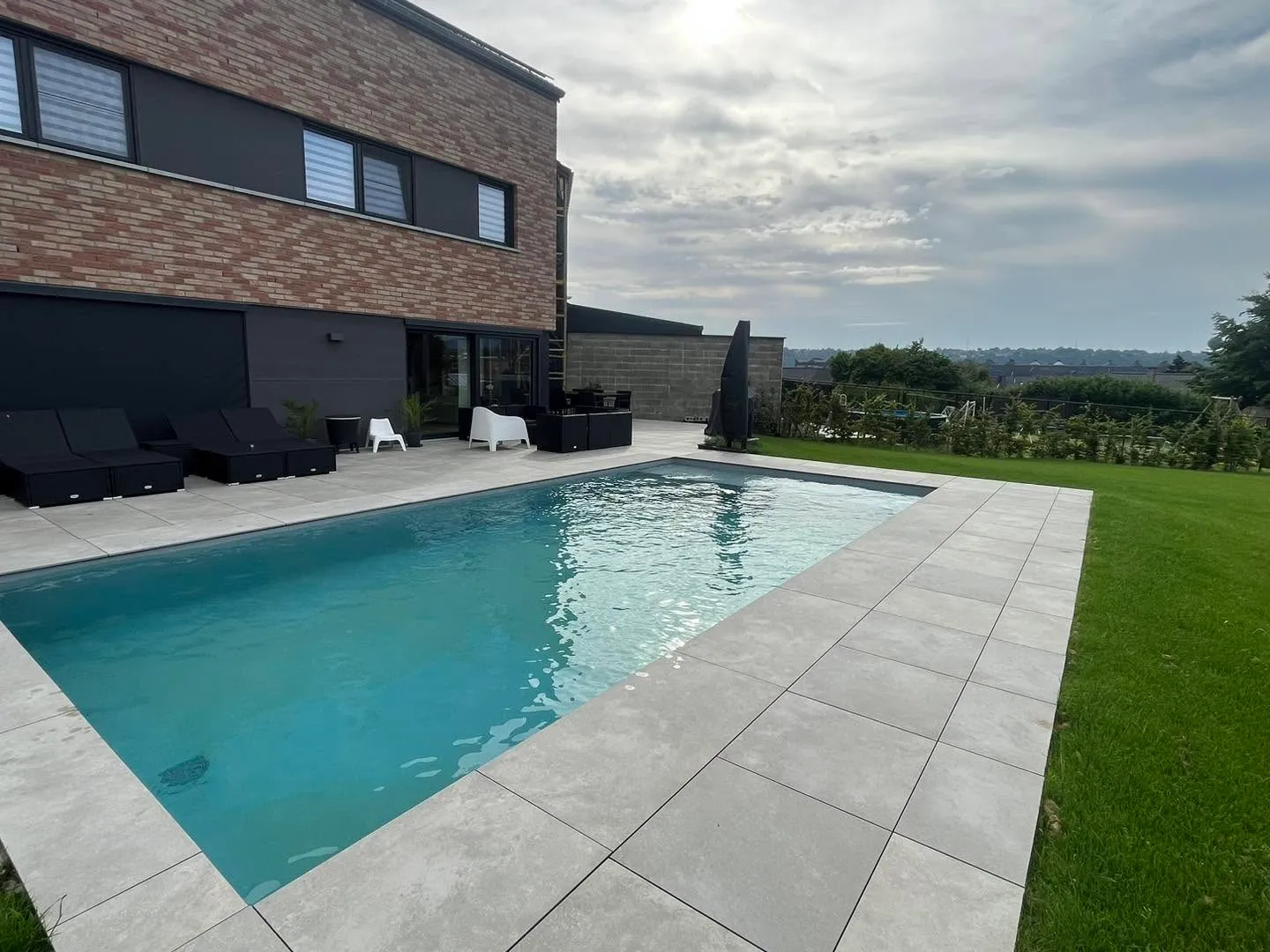 Piscine contemporaine entourée d’une terrasse claire devant une maison en briques