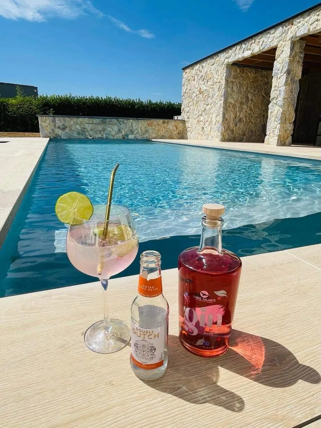 Piscine et pool house en pierre vus depuis une terrasse avec verre au premier plan